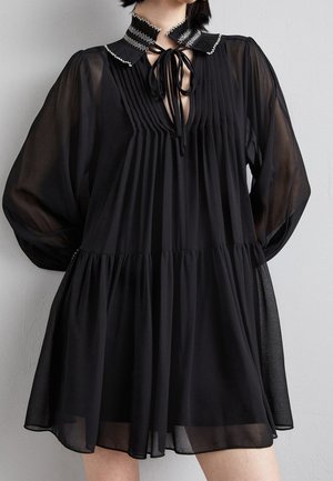 Femme portant une robe noire plissée avec des manches longues transparentes, un col volanté avec des coutures blanches et un détail noué à l'avant.