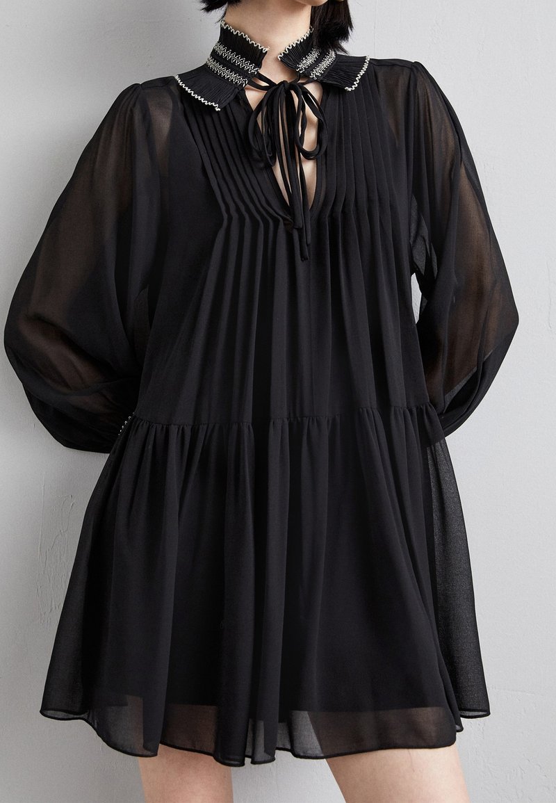 Femme portant une robe noire plissée avec des manches longues transparentes, un col volanté avec des coutures blanches et un détail noué à l'avant.