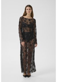 Abito in pizzo nero con maniche lunghe, caratterizzato da motivi floreali, tessuto trasparente e design lungo fino al pavimento. Indossato sopra un indumento intimo nero.