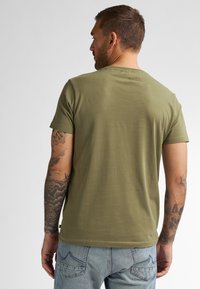 T-shirt en coton vert olive avec des manches courtes, un col rond et une texture simple et lisse. Tatouages visibles sur le bras.