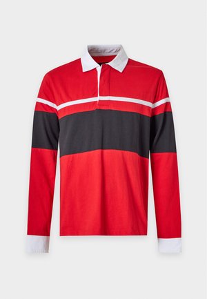 Jeune homme debout portant un maillot de rugby à manches longues rouge et noir, un jean large bleu clair et des baskets blanches sur fond blanc uni.
