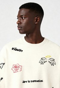 Sudadera crema con gráficos bordados, que incluyen una rosa, alas de ángel y acentos de texto en negro y rojo. Cuello redondo acanalado y textura visible.