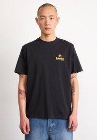 Camiseta de algodón negra con mangas cortas, cuello redondo y logo de Timberland en amarillo en el lado izquierdo del pecho; combinada con jeans de un tono azul claro.