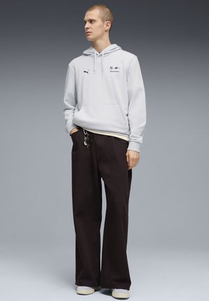 Sudadera gris con detalles de logo, bolsillo frontal y cordón en la capucha, combinada con pantalones negros de corte holgado. Zapatos claros completan el conjunto.