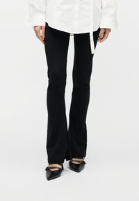 Άτομο που φορά μαύρο φαρδύ παντελόνι με εμπρόσθιες σχισμές, λευκό μακρυμάνικο πουκάμισο και μαύρα μυτερά ψηλοτάκουνα με λουράκια στον αστράγαλο.