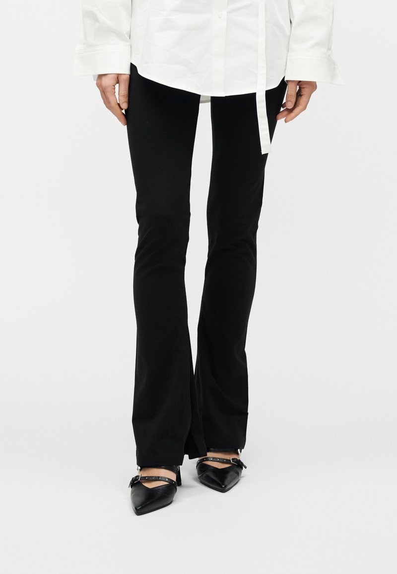 Άτομο που φορά μαύρο φαρδύ παντελόνι με εμπρόσθιες σχισμές, λευκό μακρυμάνικο πουκάμισο και μαύρα μυτερά ψηλοτάκουνα με λουράκια στον αστράγαλο.