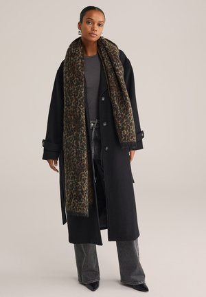 Femme portant un long manteau noir, un haut gris foncé, un jean large gris, des chaussures noires à talons pointus et une longue écharpe imprimée léopard.