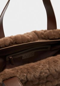Borsa a tracolla marrone con manici in pelle liscia, caratterizzata da un morbido rivestimento in pelliccia artificiale marrone e una tasca interna con zip dotata di dettagli logo.
