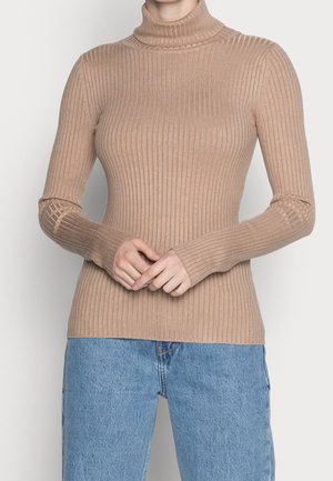 Femme portant un pull à col roulé côtelé beige ajusté et un jean bleu clair, les mains doucement jointes devant.