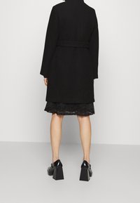 Cappotto nero lungo con vita cintura, caratterizzato da una silhouette strutturata, abbinato a un vestito di pizzo nero a strati e scarpe con tacco spesso.