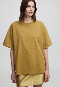 T-shirt en coton jaune à manches courtes, avec des anneaux en métal décoratifs cousus dans le tissu et une coupe décontractée.