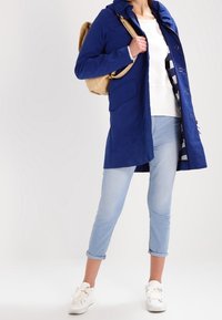 Blå, lättvikts, huva jacka med sidofickor, knäppning och kontrasterande innerfoder, bärs över en vit topp och ljus jeans.