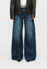 Jeans in denim a gamba larga di colore blu scuro, con un design classico, lievi sfumature e orlo grezzo. Abbinati a una giacca di pelle nera e una cintura.