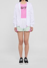 Bílá lehká bunda, růžové tričko se třpytivým logem, šortky s mint a růžovým gradientem a černé sportovní boty s texturovanými detaily.