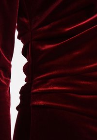 Tissu en velours bordeaux avec une texture lisse, présentant des fronces sur les côtés et un design ajusté, mettant en valeur une couleur riche et un éclat subtil.