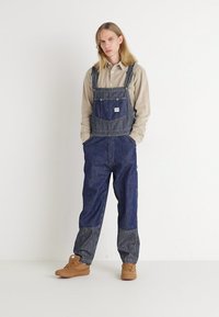 Pantalones de mezclilla estilo overall con un diseño de rayas azules y blancas. Incluye un bolsillo delantero, corte holgado y dobladillos acampanados. Combinados con una camiseta beige y zapatos marrones.