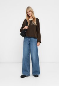 Suéter oversized castanho escuro com mangas largas, usado com jeans de perna larga azul clara. Pequena mala preta de ombro e sapatos pretos de deslizar.