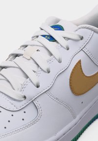 Λευκό αθλητικό παπούτσι Nike με ταμπά σήμα Swoosh, λευκά κορδόνια και μπλε-πράσινο κυκλικό λογότυπο στη γλώσσα.