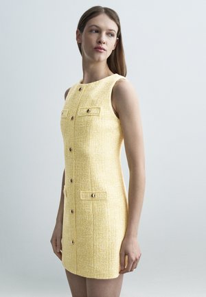 Femme portant une robe jaune texturée sans manches avec boutons à l'avant et poches, debout devant un fond gris clair uni.