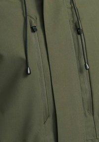 Chaqueta verde oliva hecha de tejido texturizado, con dos bolsillos con cremallera, cordones ajustables y acentos de metal negro en contraste.