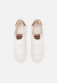 White leather sneakers with brown leather heel accents; features flat white laces and a branded tab on the tongue. Smooth texture throughout.