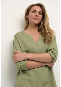 Zelena lanena srajca z V-izrezom, zavihki na rokavih in subtilne šivane detajle, nosi manekenka z prekriženimi rokami pred nevtralno ozadje.