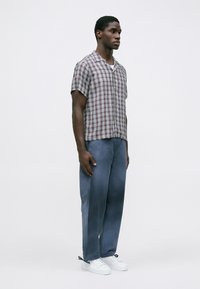 Uomo in piedi con pantaloni blu, sneakers bianche, camicia a quadri a manica corta con bottoni, su uno sfondo chiaro e uniforme.