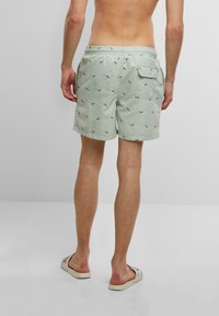 Svijetlozelene kupaće hlače s mekom teksturom, koje sadrže uzorak morskog psa u tamnoplavoj boji. Imaju elastičnu traku u struku i stražnji džep.