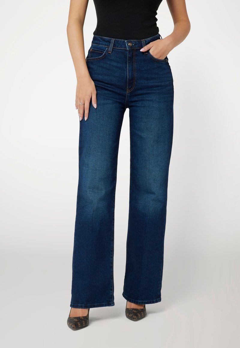 Mörkblå högmidjade jeans med vida ben, som har fem fickor och kontrasterande sömmar, tillsammans med svarta klackar.