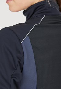 Svart och marinblå jacka med hög krage, slät textur och kontrasterande paneler med tunna grå detaljer längs sömmarna.