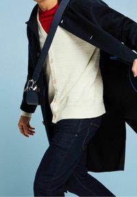 Personne portant un manteau bleu foncé, un cardigan blanc boutonné, une chemise rouge, un jean foncé et un petit sac bandoulière bleu marine sur un fond bleu.