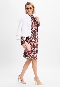 Bloemenpatroon jurk in roze en bruin, gecombineerd met een witte denimjas, beige hakken en een lichtroze clutch. Ronde, roze zonnebril inbegrepen.