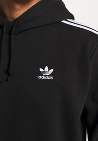 Schwarzer Baumwoll-Hoodie mit einem weißen bestickten Adidas-Logo auf der Brust und dem klassischen weißen Drei-Streifen-Design entlang der Schultern.