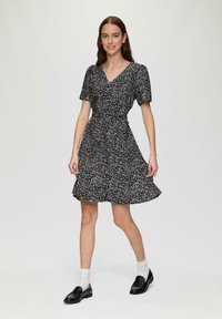 Schwarzes Kurzarmkleid mit floralem Muster, V-Ausschnitt, tailliertem Schnitt und schwingendem Rock, kombiniert mit schwarzen Mokassins und weißen Socken.