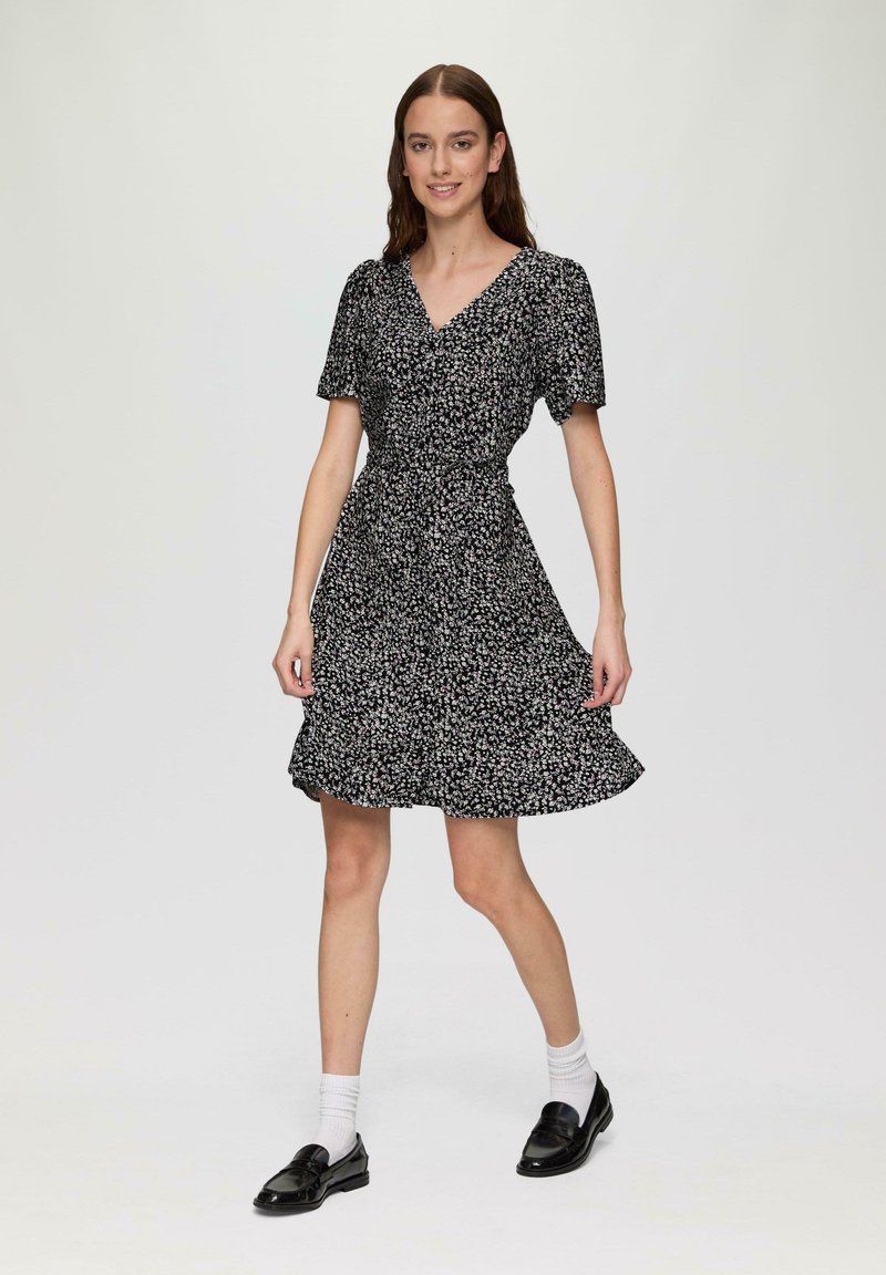 Schwarzes Kurzarmkleid mit floralem Muster, V-Ausschnitt, tailliertem Schnitt und schwingendem Rock, kombiniert mit schwarzen Mokassins und weißen Socken.