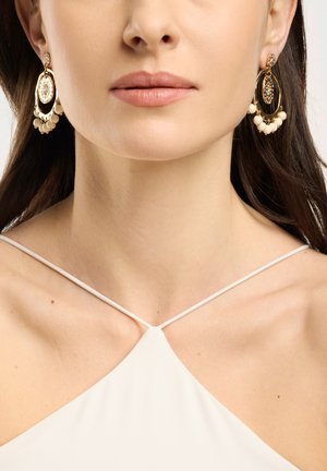 Kvinde iført gyldne hoop-øreringe med cremefarvede perleaccents og en hvid haltertop med tynde stropper.