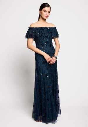 Femme debout portant une robe de soirée bleu marine épaules dénudées avec des ornements en sequins floraux et une jupe transparente superposée, mains jointes devant.