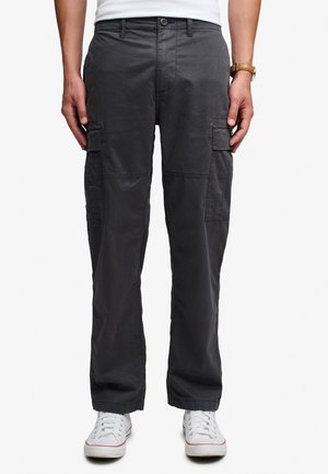 Homme portant un pantalon cargo gris foncé avec des poches latérales, des baskets blanches et un haut blanc, debout devant un fond uni.