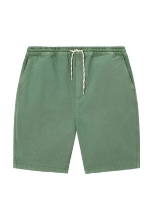 Homme debout portant un sweat-shirt marron, un pull jaune drapé sur les épaules, un short vert, des chaussettes vertes à motifs et des chaussures beige.