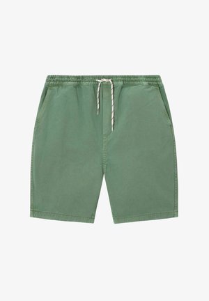 Mann steht, trägt einen braunen Sweatshirt, einen gelben Pullover über den Schultern, grüne Shorts, gemusterte grüne Socken und sandfarbene Schuhe.