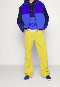 Chaleco de forro polar azul sobre sudadera de capucha azul con estampado, combinado con pantalones de pana amarillos y zapatillas de deporte azul marino. Se presentan tejidos texturizados y colores vibrantes.