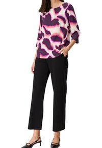 Bedruckte Bluse mit lila, rosa und weißen Mustern, kombiniert mit schwarzen Hosen. Die Bluse hat einen runden Halsausschnitt und drei Viertelärmel.