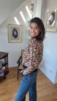 Frau lächelt über die Schulter, trägt ein florales Oberteil und blaue Jeans in einem Raum mit abstrakter blauer Kreis-Kunst und Holzmöbeln.