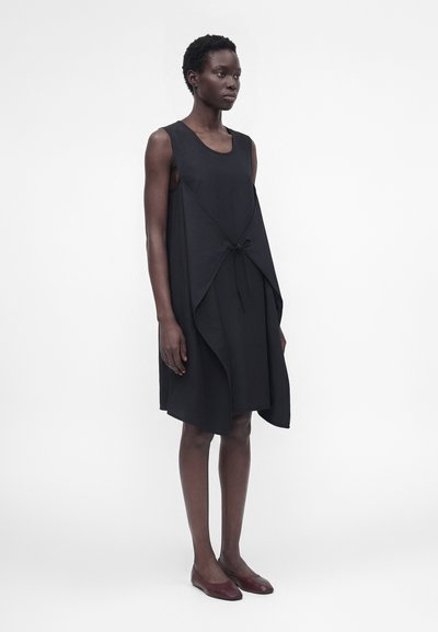 Ψηλή γυναίκα με κοντά σγουρά μαλλιά φοράει μαύρο αμάνικο φόρεμα με δεμένο επίχρισμα, συνδυασμένο με μπορντό πλατφόρμες, στέκεται μπροστά σε λευκό φόντο.