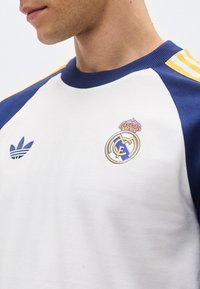 Homme portant un maillot de sport blanc et bleu avec les logos Adidas et Real Madrid sur la poitrine et des bandes jaunes sur l'épaule.