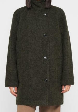 Personne portant un manteau en laine texturée vert olive foncé avec un col haut et quatre boutons noirs, associé à un pantalon beige.