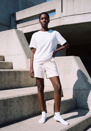 Une personne portant un tee-shirt blanc, un short et des chaussettes pose sur des marches en béton devant un bâtiment moderne.