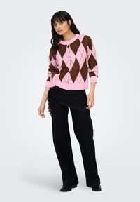 Rosa-brauner, argylemusternder Cardigan mit rundem Ausschnitt, Knopfverschluss und langen Ärmeln, kombiniert mit schwarzen, strukturierten, weit geschnittenen Hosen.