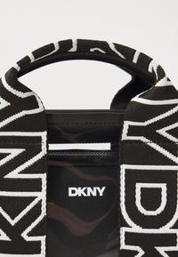 Czarna torebka na ramię z teksturowanym, błyszczącym wykończeniem, z białymi detalami logo DKNY na plecionym pasku. Udekorowana odważnymi wzorami.