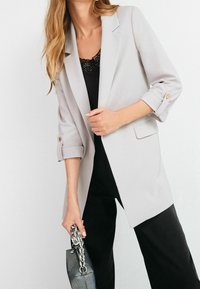 Blazer gris clair avec des manches retroussées, comportant des boutons dorés. Associé à un haut noir et tenant un sac à main avec des détails en chaîne argentés.
